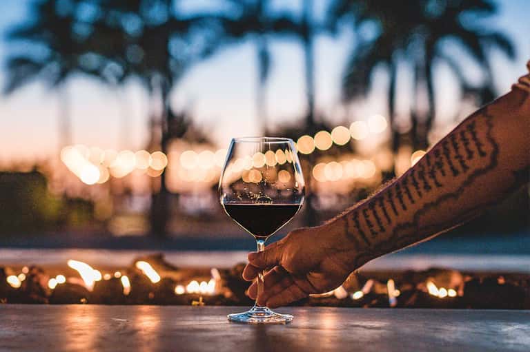 Hand holding wine glass at sunset with palm trees and string lights in background