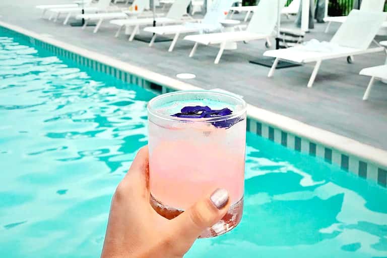 Hand holding pink cocktail with blue orchid garnish by turquoise pool with white lounge chairs