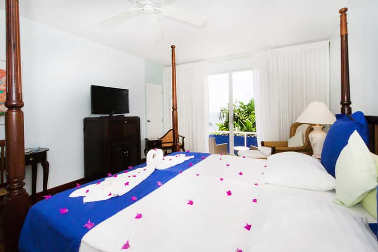 Resort bedroom with four-poster wood bed, blue decorative runner with pink flowers, ocean view from balcony.