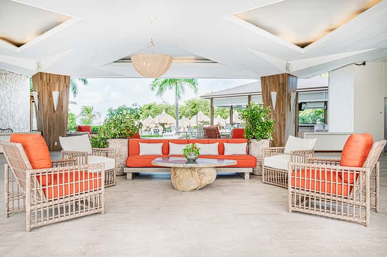 Covered resort lounge with white and orange woven chairs, coral cushions, and tropical greenery