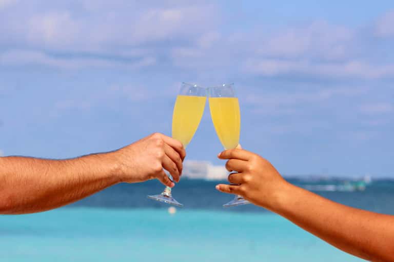 Two hands toasting champagne glasses with turquoise ocean and sailboats in background