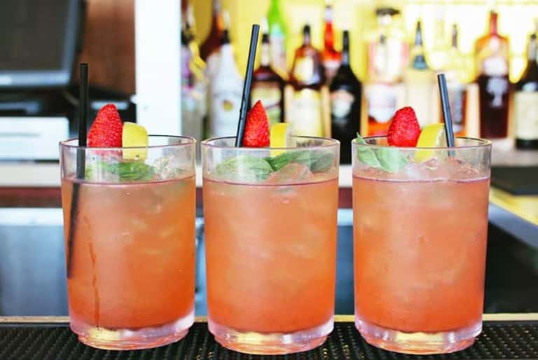 Three coral-colored cocktails with strawberry and lime garnishes and blue straws on a bar counter