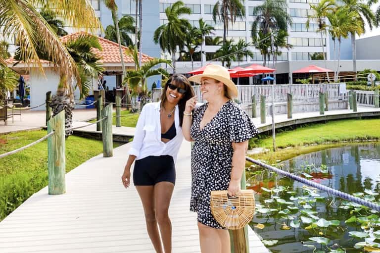 Two women walking on wooden boardwalk beside marsh area with palm trees and hotel buildings in background