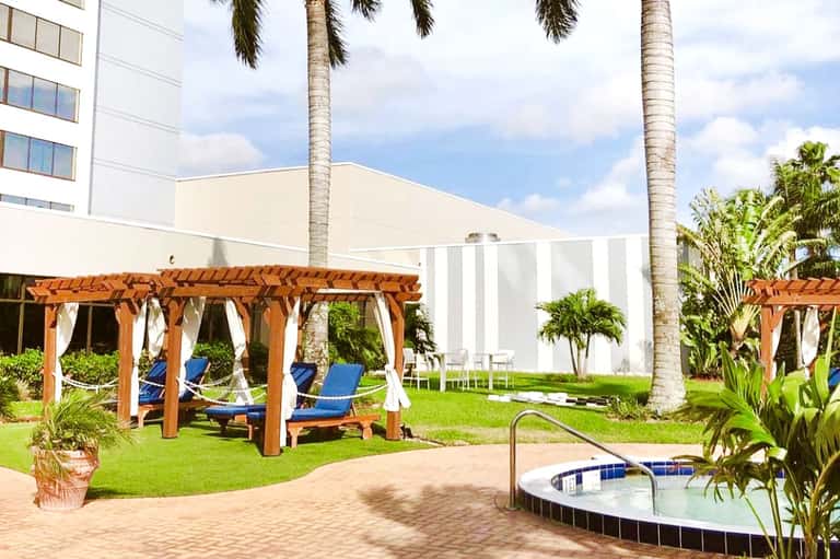Resort courtyard with wooden pergolas, blue lounge chairs, tropical plants, and hot tub on manicured lawn
