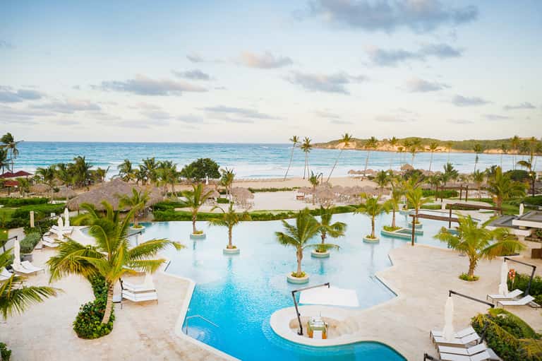 Tropical resort pool with curved design, palm trees, white loungers, and ocean beach in background