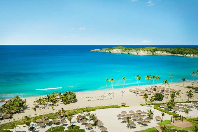 Aerial view of pristine turquoise beach with palm umbrellas, palm trees, and island cliff backdrop