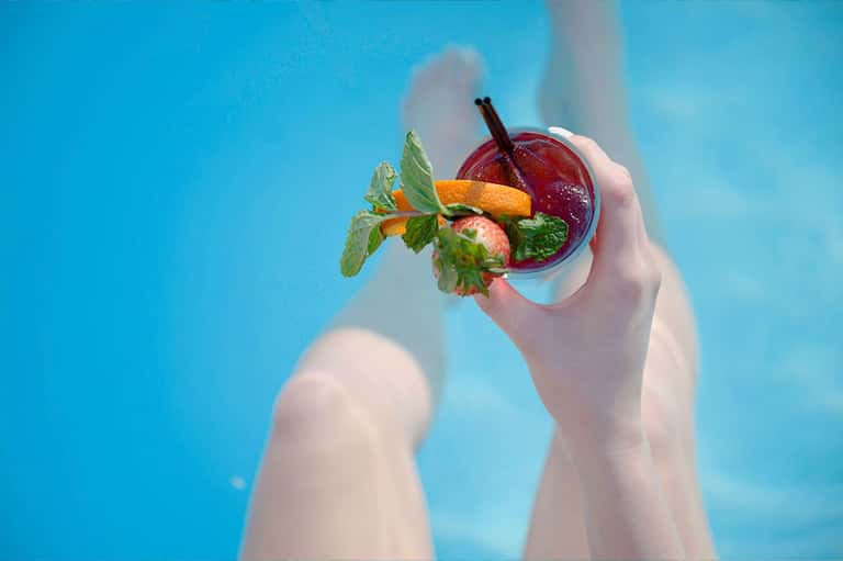 Hands holding tropical cocktail with fresh mint and fruit over blue pool water