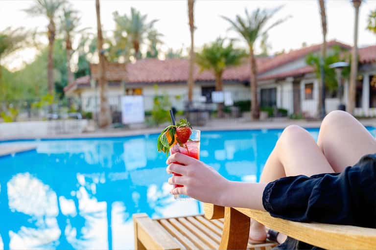 Person holding tropical cocktail overlooking resort pool and palm trees