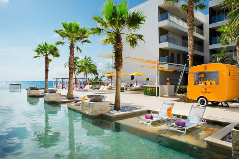Beach resort pool area with palm trees, loungers, orange umbrellas, and beverage station
