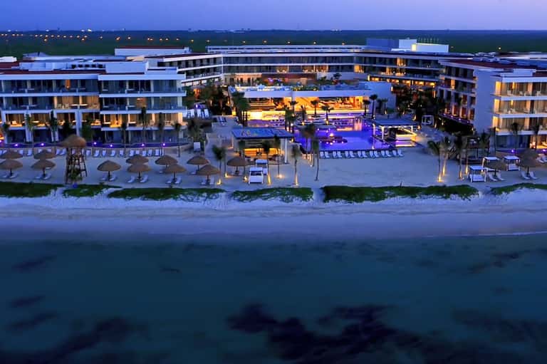 Beachfront resort at dusk with illuminated pools, loungers, umbrellas, and modern curved architecture