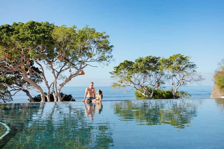 Man and woman relaxing in an infinity pool with trees and ocean horizon views