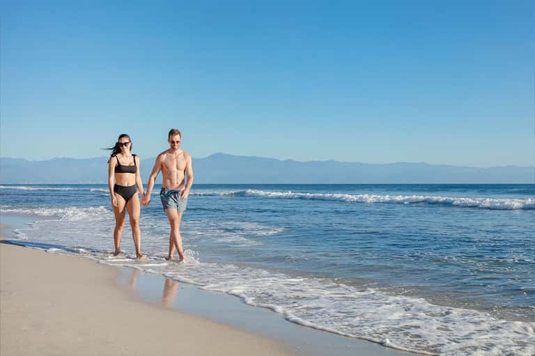 Couple wading in ocean surf on sandy beach with mountains visible in distance