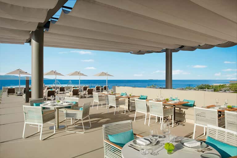 Coastal dining terrace with white furniture, turquoise accents, umbrellas, and ocean views