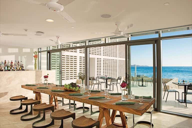 Modern restaurant interior with wooden dining tables, ocean views, and minimalist décor