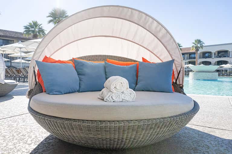 Woven daybed with blue pillows and white canopy overlooking a resort pool and palm trees