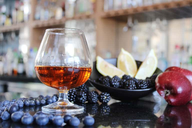 Brandy snifter with dark liquor surrounded by berries, citrus, and pomegranate at bar