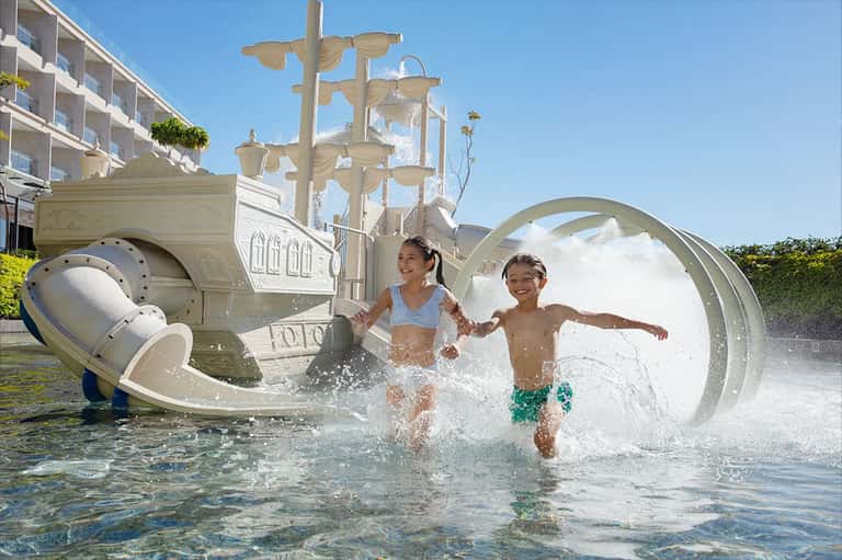 Children playing on elaborate ship-themed splash pad with water features and waterslides