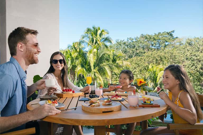 Family enjoying breakfast on a terrace with pool views, palm trees, and green landscaping