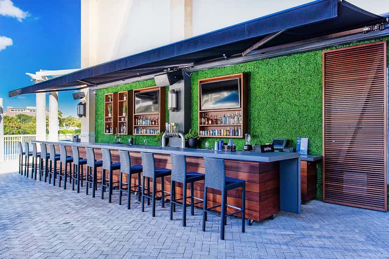 Outdoor bar with green wall, dark seating, and blue striped canopy overlooking resort grounds