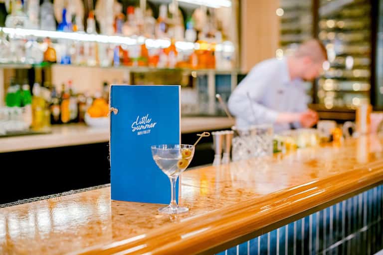 Hotel bar counter with blue menu holder, cocktail glass, and bartender in background with liquor bottles