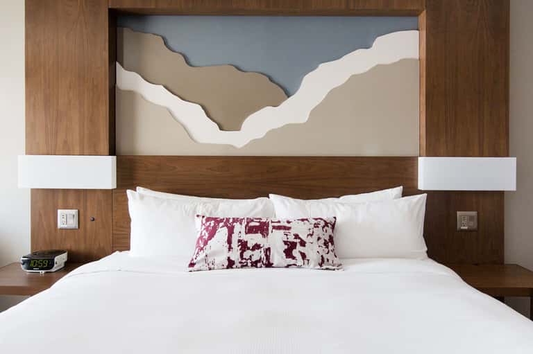 Modern hotel bedroom with wooden headboard, white bedding, burgundy accent pillow, and minimalist artwork