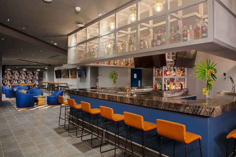Modern hotel bar with blue counter, orange seating, and illuminated bottle display