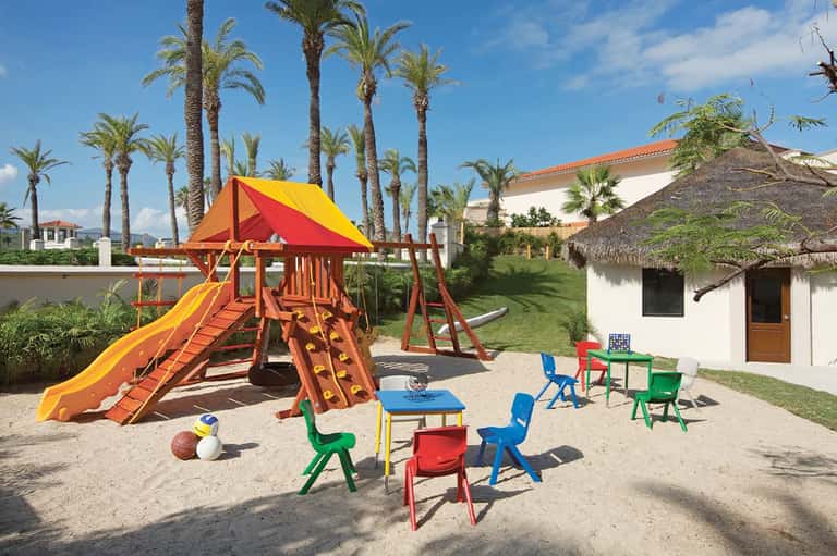 Colorful children's playground with red and yellow play structure surrounded by palm trees and resort grounds