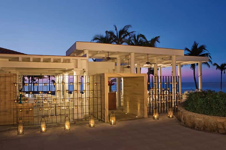 Modern beachfront structure with glass walls, white columns, and palm trees at dusk