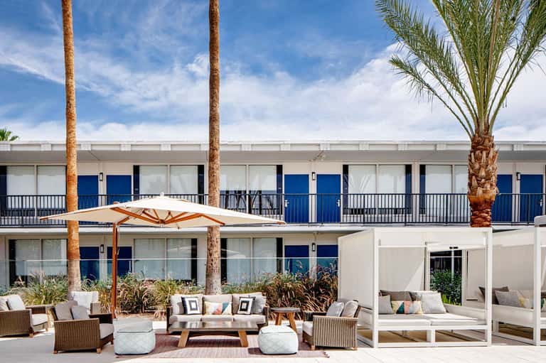 Modern hotel courtyard with lounge seating, palm trees, orange umbrella, and two-story building with blue doors