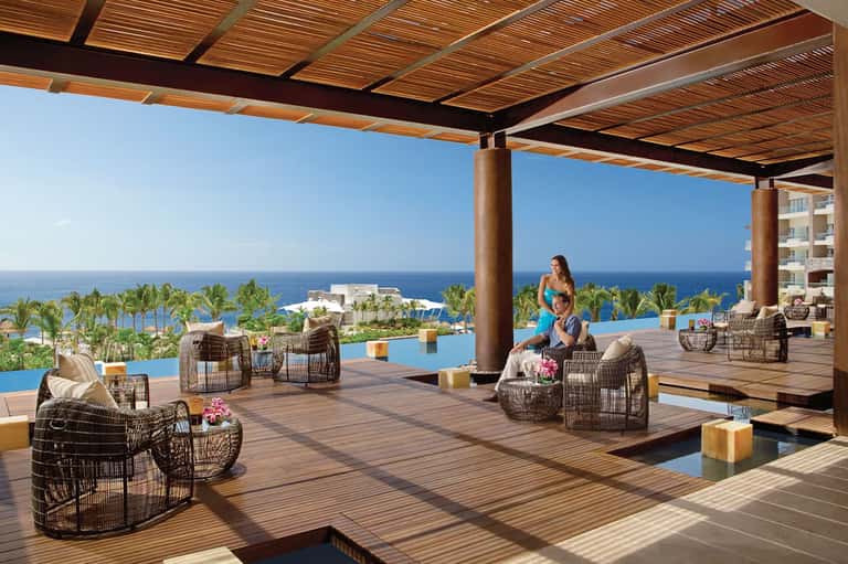 Oceanfront lounge with woven chairs, wooden pergola, infinity pool, and panoramic sea views