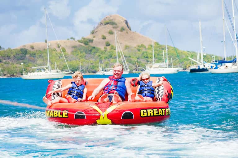 Children on red inflatable tube being pulled in turquoise water near sailboats