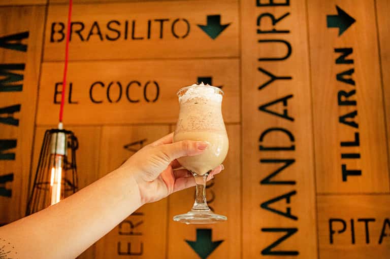 Hand holding creamy tropical cocktail against wooden menu board with drink names
