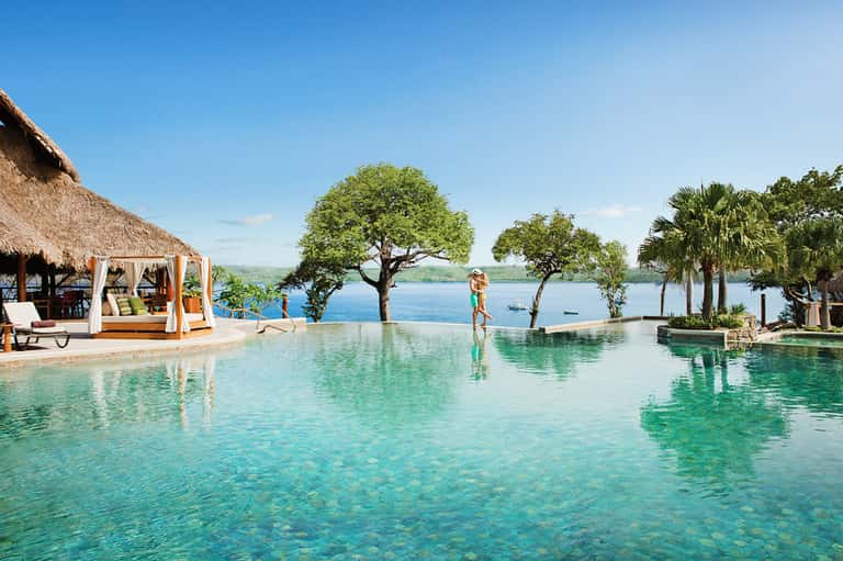 Infinity pool overlooking tropical lake with thatched cabanas, palm trees, and mountain views