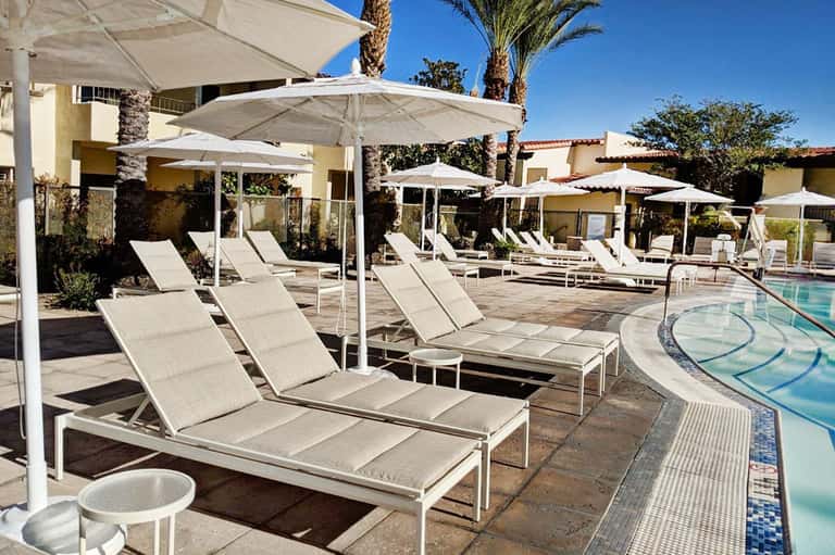 Resort pool deck with white umbrellas, lounge chairs, and palm trees under clear blue sky