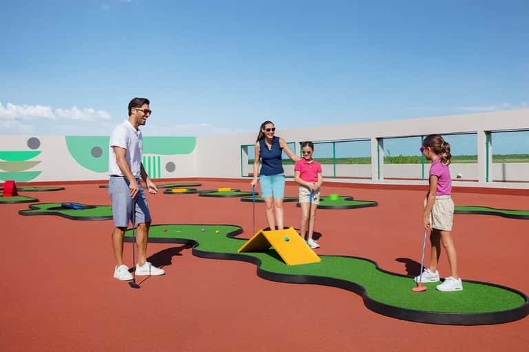 Family playing miniature golf on colorful course with red surface and green obstacles