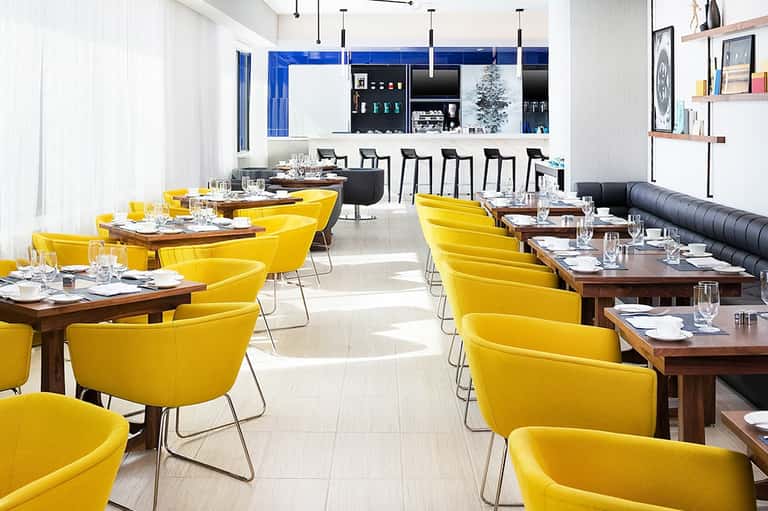 Modern restaurant dining room with yellow chairs, wooden tables, bar seating, and blue accent wall