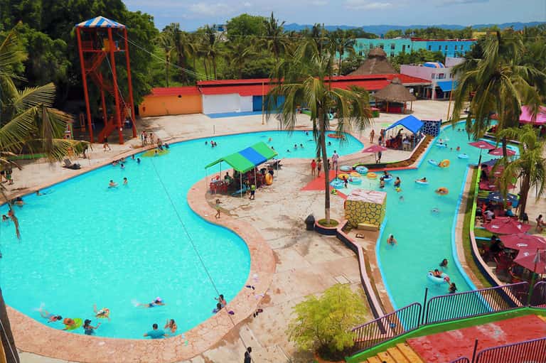 Large turquoise community pool with palm trees, water slide, and colorful resort buildings