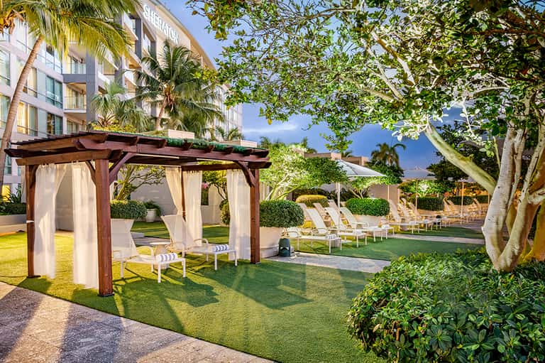 Resort lawn area with wooden pergola, cabanas, white curtains, and mature trees