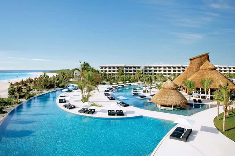 Large resort pool complex with thatched structures, white loungers, and beachfront location