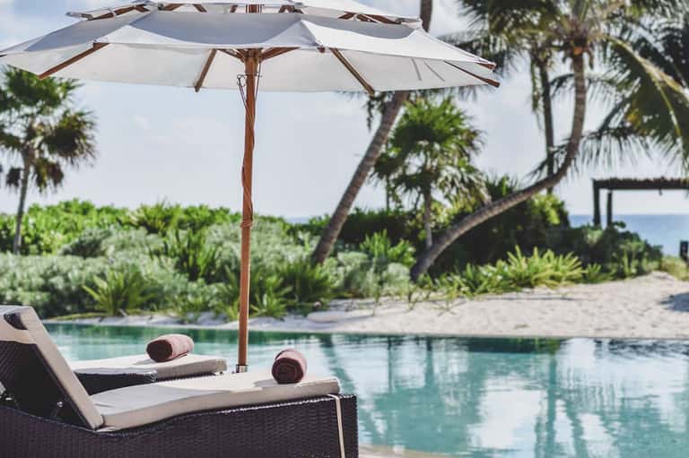 Luxury poolside lounge chairs under wooden umbrella with ocean view and palm trees