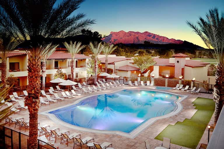 Resort pool at dusk with illuminated blue water, palm trees, lounge chairs, and red mountain backdrop