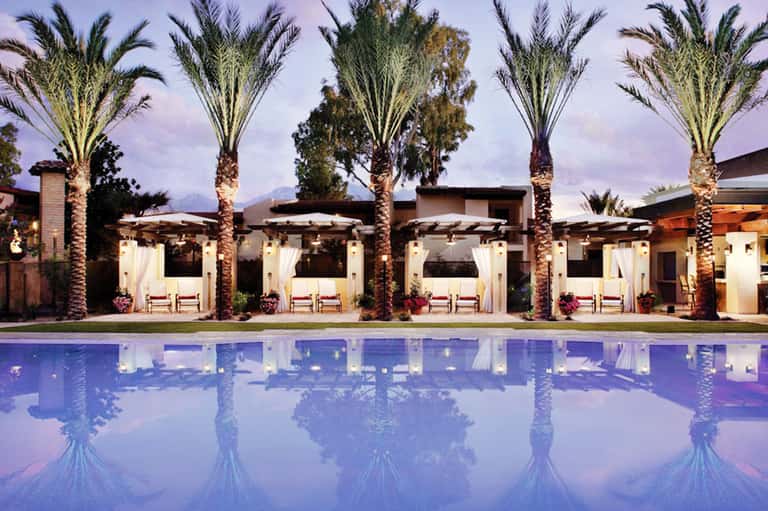 Elegant poolside cabanas with white curtains and palm trees reflected in still water at dusk