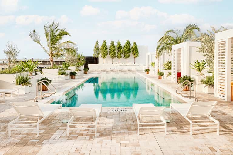 Modern resort pool with white lounge chairs, tropical landscaping, and buildings