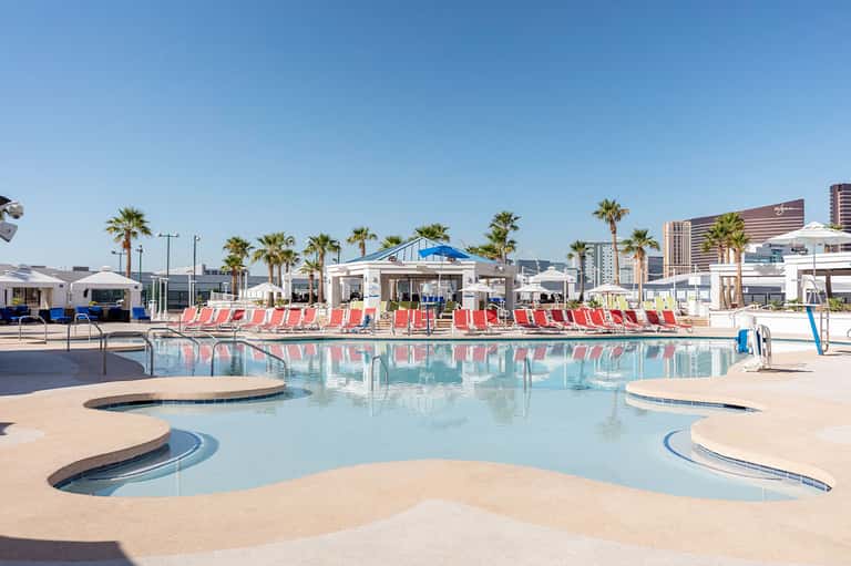 Resort pool with red lounge chairs, palm trees, and high-rise buildings in background