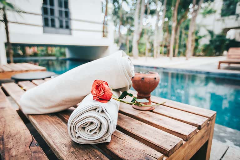 Rolled white spa towels with red rose on wooden deck overlooking illuminated resort pool