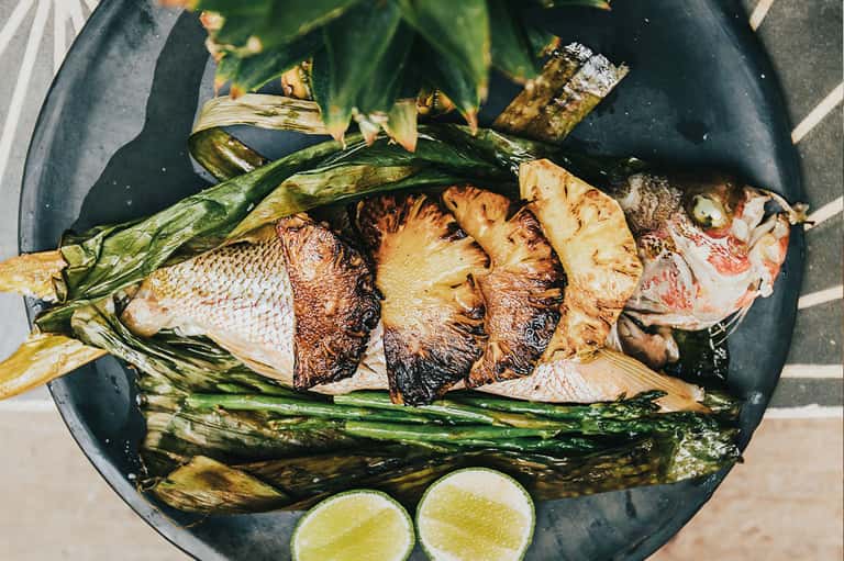 Grilled whole fish with charred vegetables and lime on dark plate, top view
