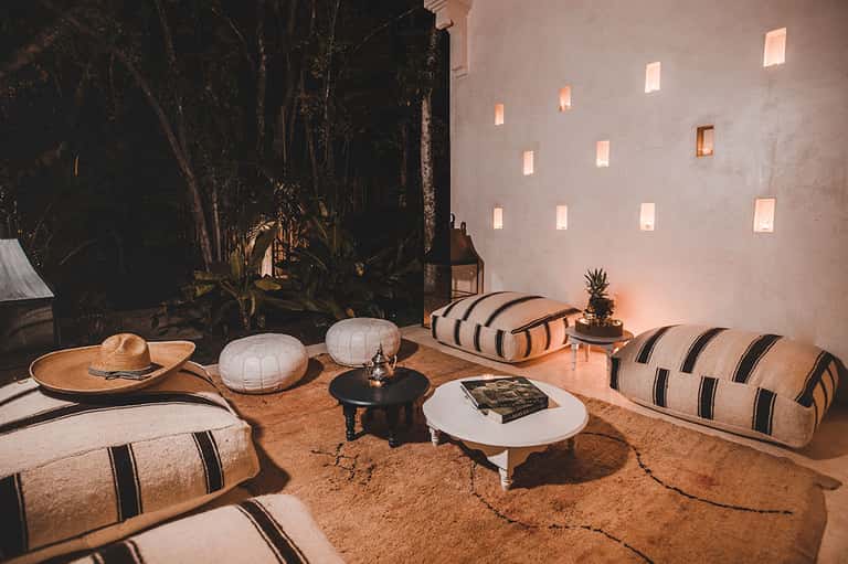 Bohemian outdoor lounge with white and striped poufs, dark wooden wall, ambient evening lighting