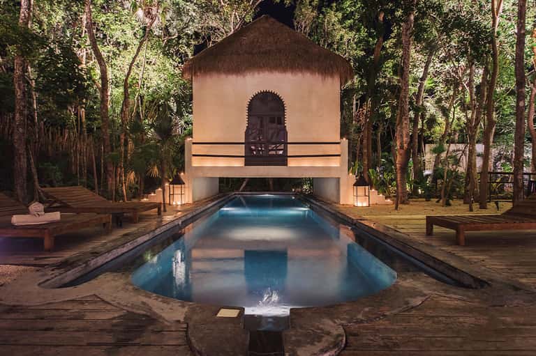 Illuminated swimming pool at night beneath Moroccan-style pavilion in jungle setting