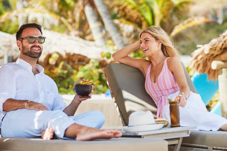 Couple relaxing on beach chairs with tropical palm trees, smiling and holding cocktails