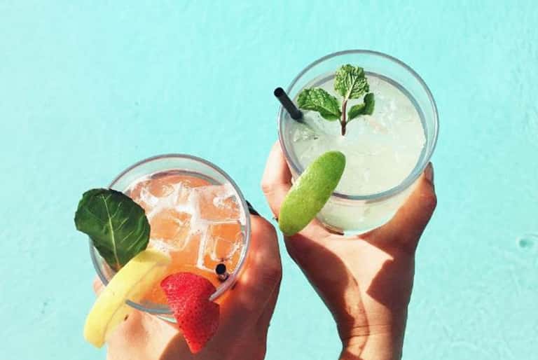 Two tropical cocktails with fresh fruit garnishes and mint leaves held against a turquoise background